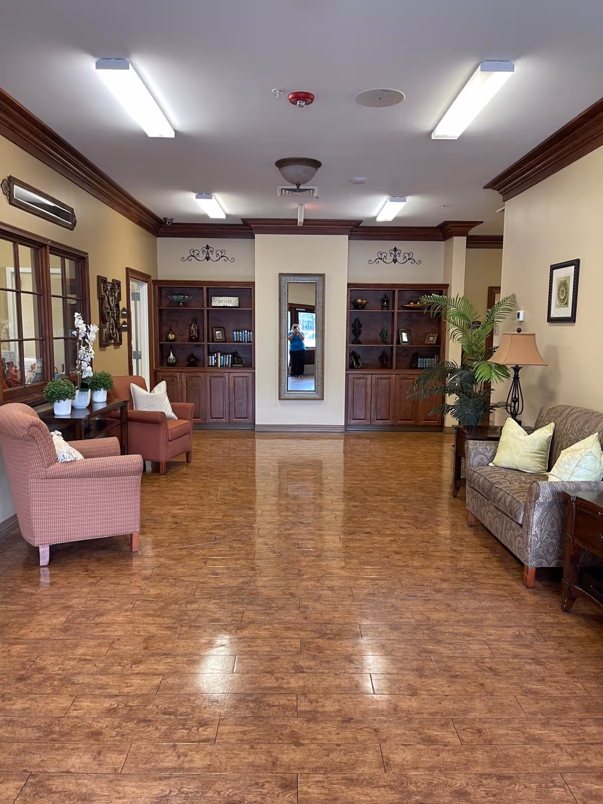A spacious, well-lit living room area with wooden flooring, two upholstered armchairs on the left, a patterned sofa with cushions on the right, a side table with a lamp, and built-in wooden shelves with decorative items and books on the far wall. A large mirror is centered between the shelves, reflecting a person taking the photo. There are plants and framed artwork on the walls.