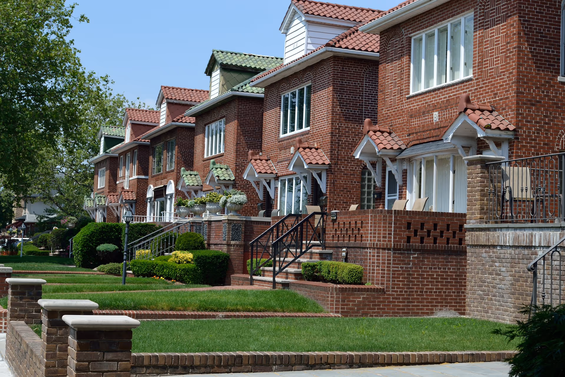 Row of red-brick townhouses with tiled roofs, front steps, and manicured lawns.