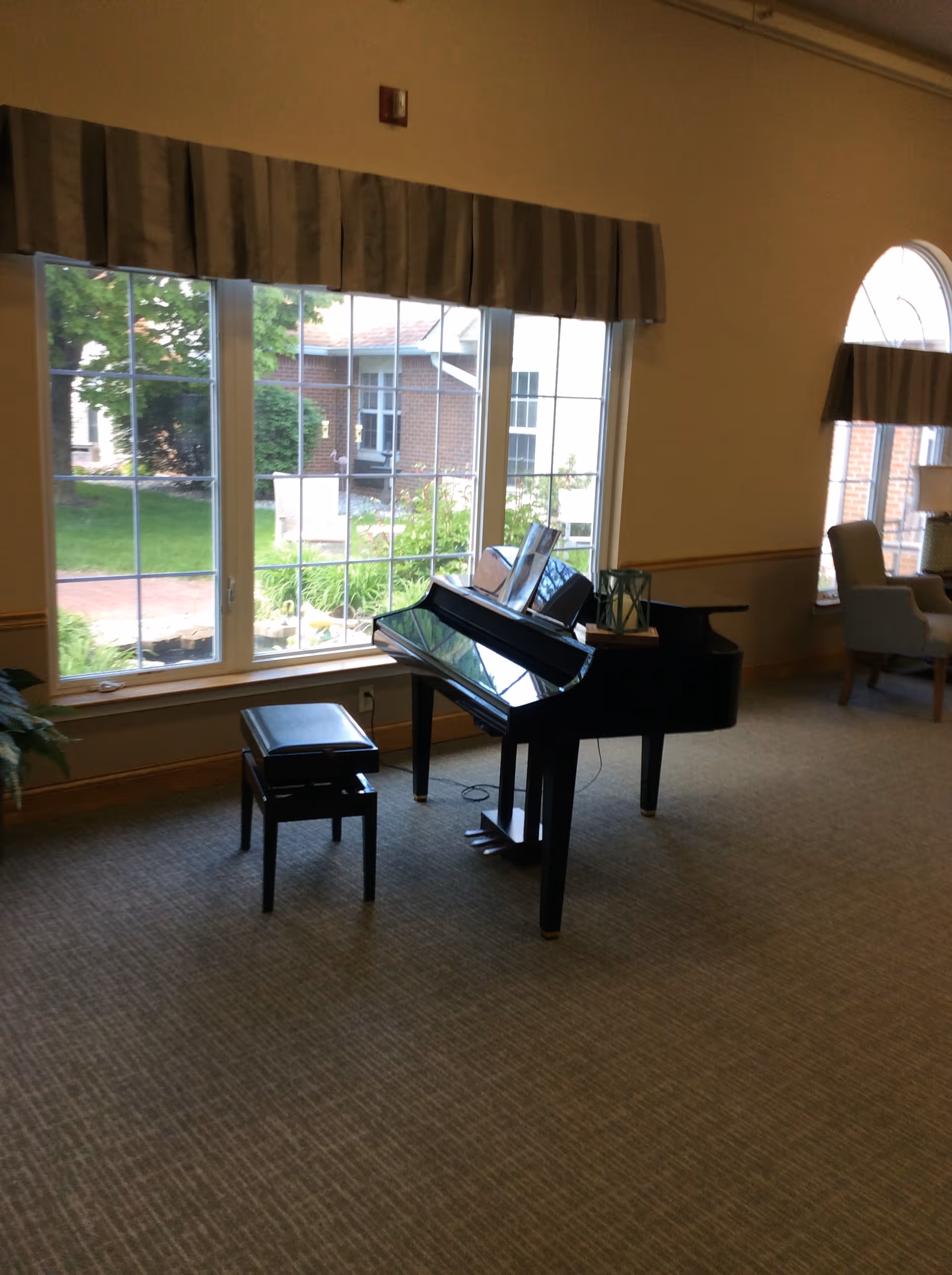 Interior room with a black grand piano and matching bench placed near large windows with a view of a garden outside. The room has beige carpet, light-colored walls with a chair rail, and a seating area with armchairs near another window with an arched top.