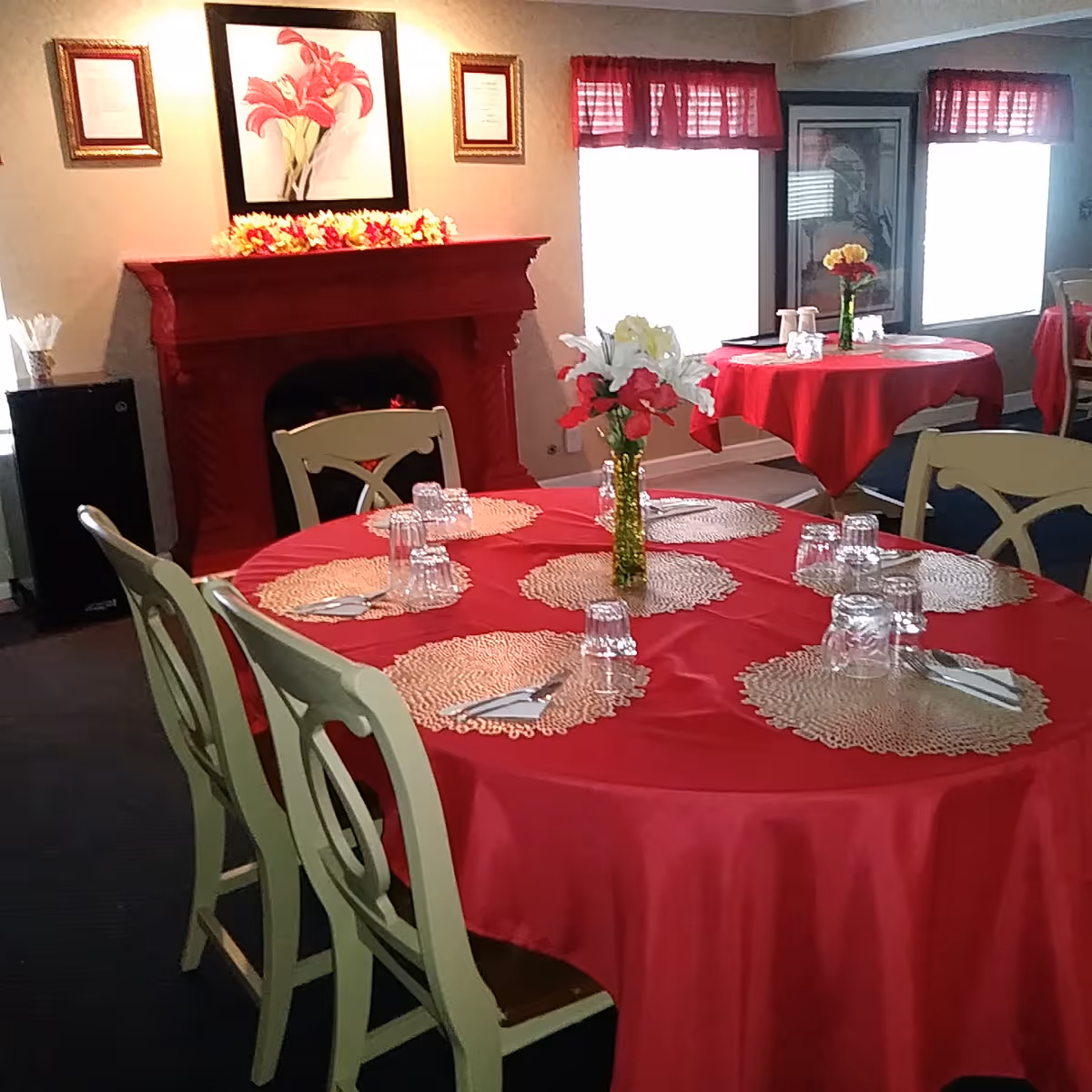 Dining room with round tables covered in red tablecloths, place settings and floral centerpieces, and a decorative red fireplace in the background.
