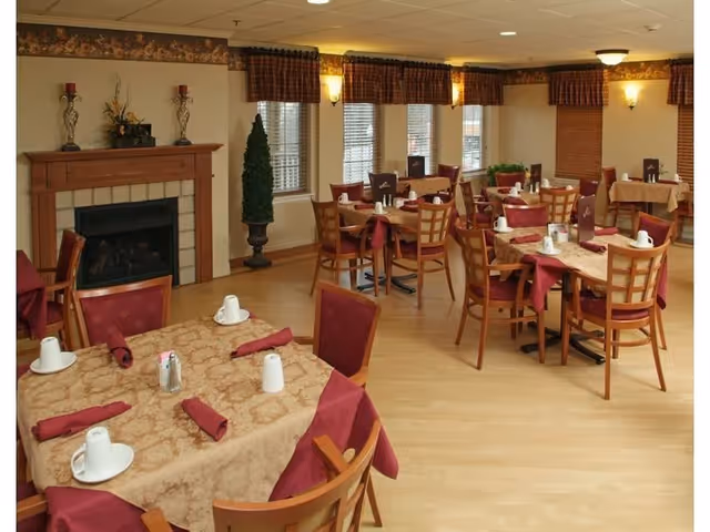 Well-lit senior dining room with multiple tables set with cups and napkins, wooden chairs, and a fireplace.