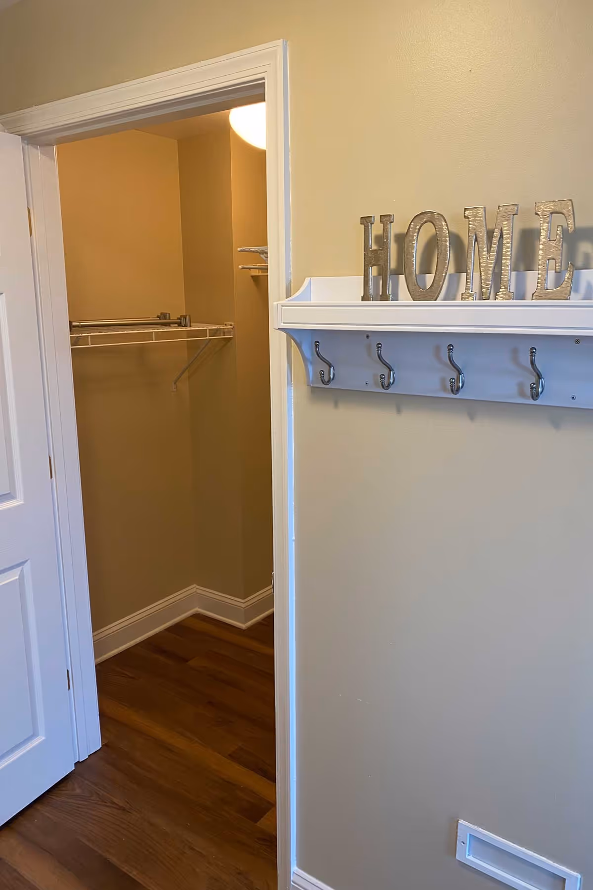 Interior view showing a wall-mounted white coat rack with decorative letters spelling HOME and an open door to a small walk-in closet with shelving and wood floors.