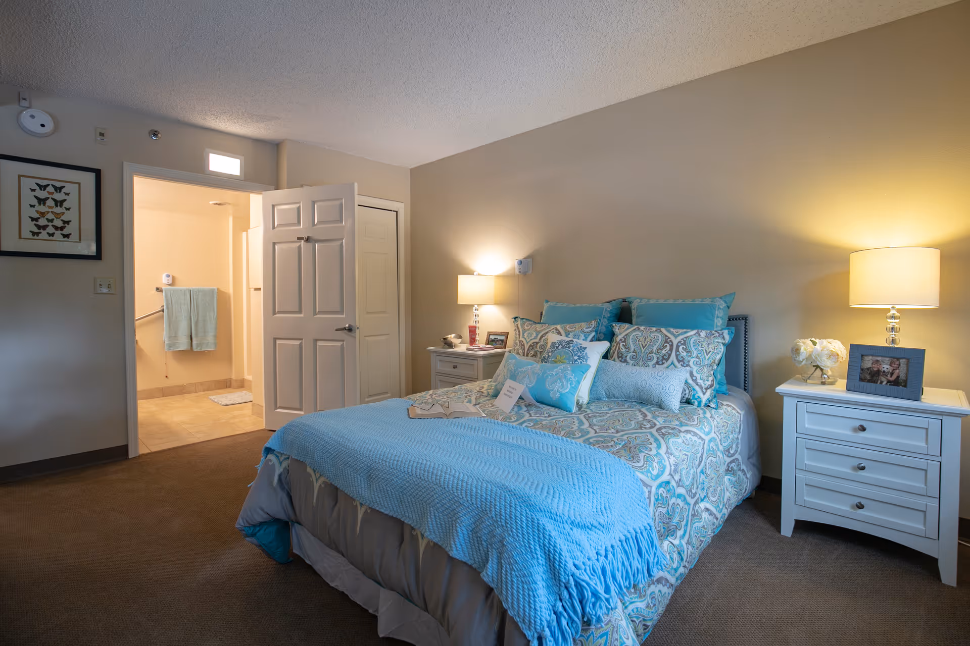 A cozy bedroom with a neatly made bed featuring blue and patterned bedding. Two white nightstands flank the bed, each with a lamp and decorative items. The door to an adjoining bathroom is open, showing a towel rack with two green towels and tiled flooring. A framed butterfly artwork hangs on the wall near the bathroom door.