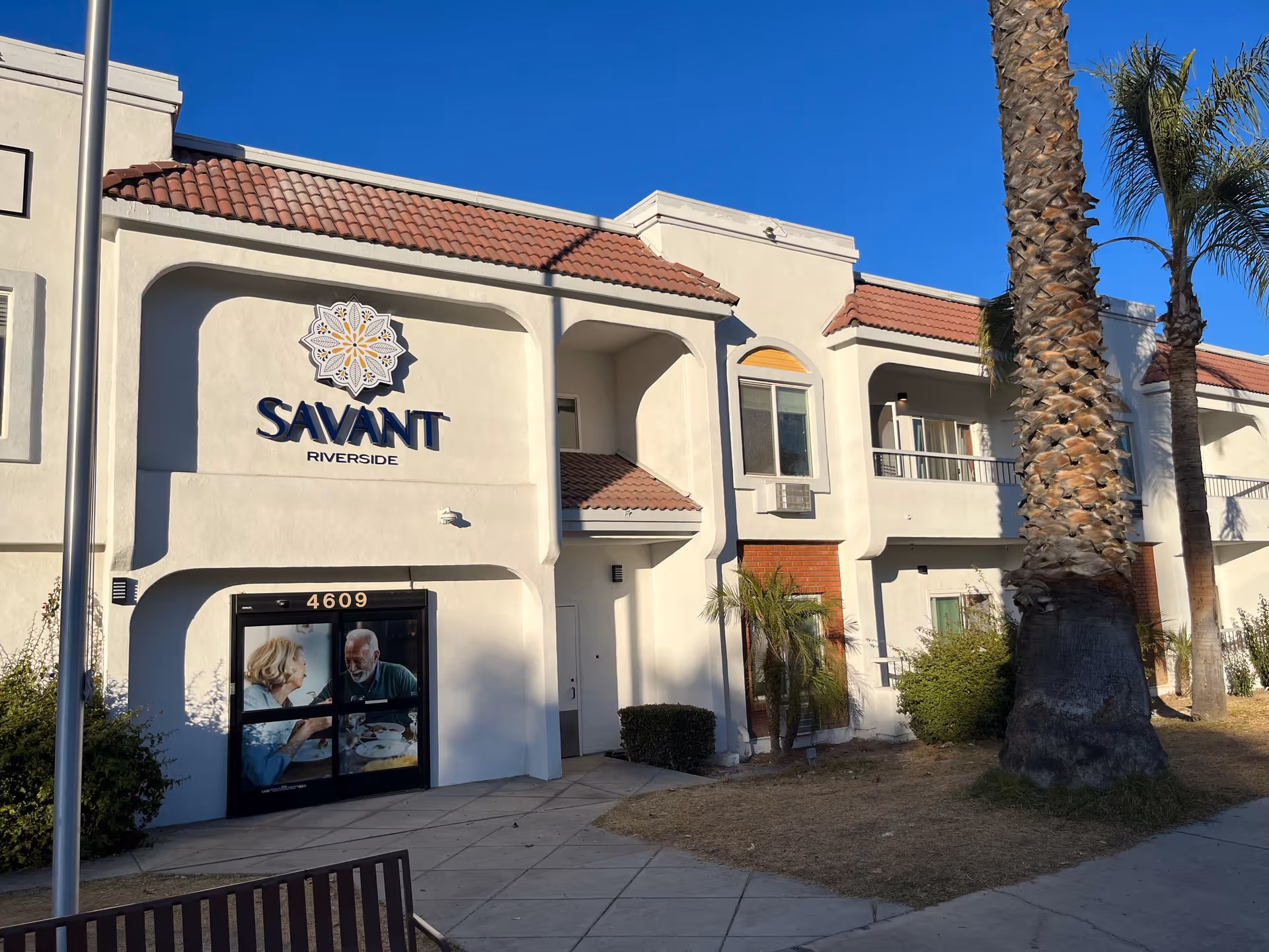 Exterior view of Savant of Riverside senior living facility building with white walls, red tile roof, palm trees, and a sidewalk in front. The entrance door has an image of an elderly man and woman dining together.