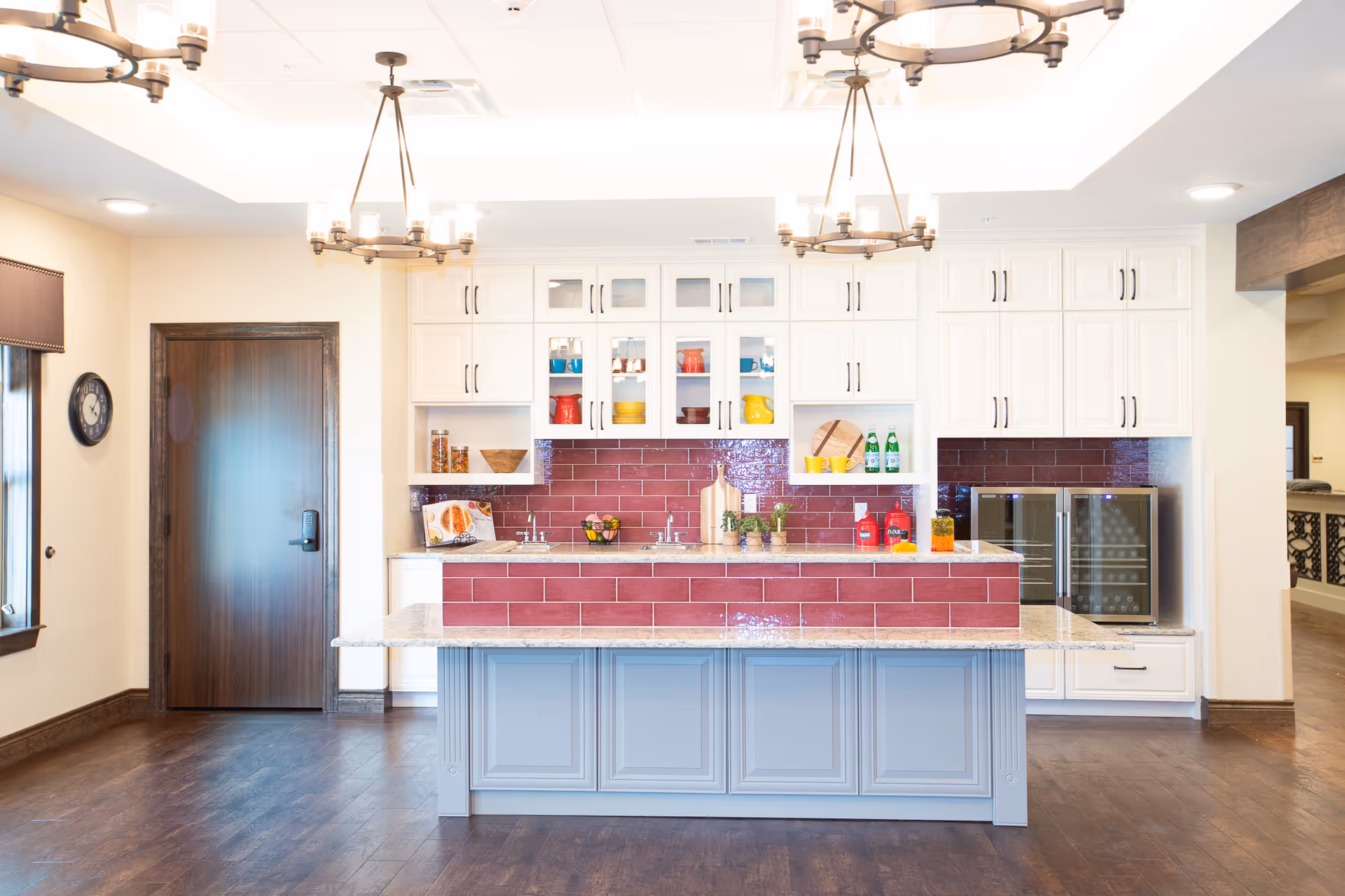 Bright and modern kitchen area with white cabinets, a large island with a gray base and granite countertop, and a red tiled backsplash. The kitchen features two chandeliers, a wooden door, a clock on the wall, and various kitchen items such as colorful bowls, bottles, and plants on the counters and shelves.