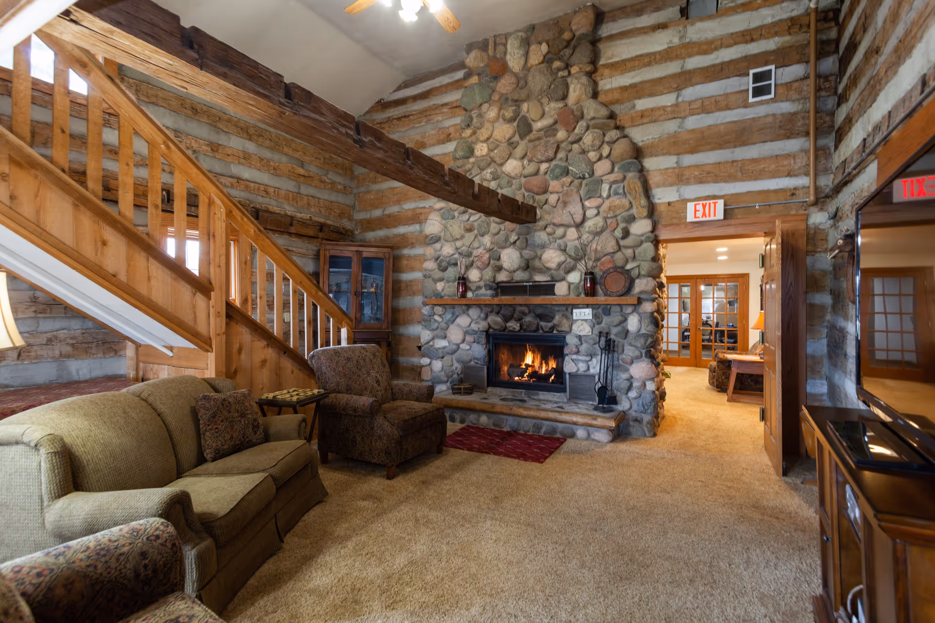 Rustic living room with a large stone fireplace, upholstered sofas, an armchair, and a wooden staircase under exposed beams.