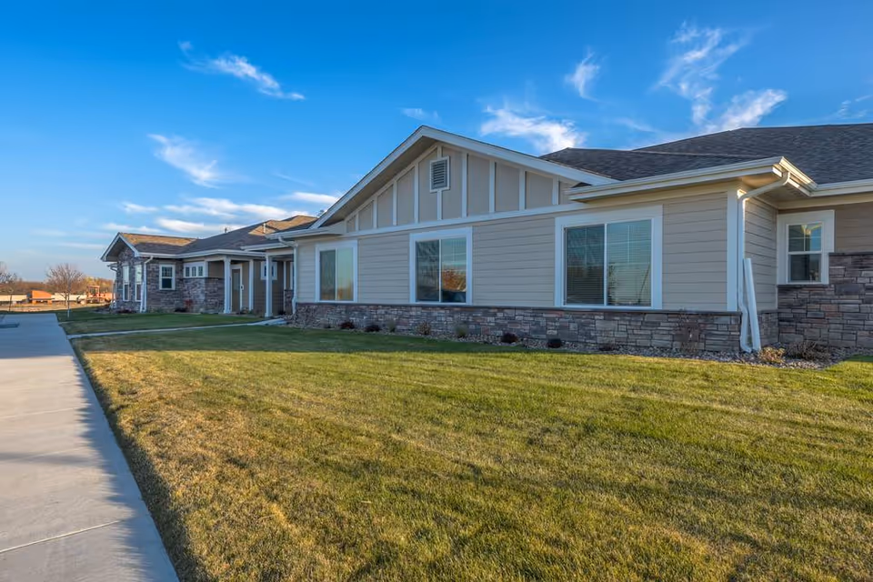 Exterior view of a single-story senior living facility with beige siding, stone accents, and a well-kept lawn under a clear blue sky.
