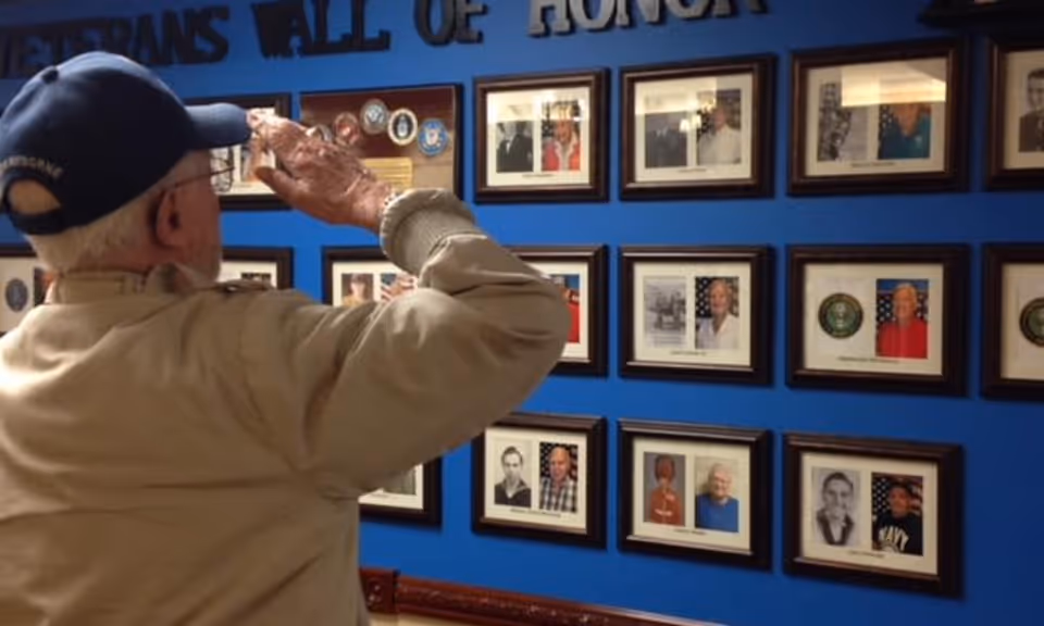 An elderly man wearing a cap and beige jacket salutes a Veterans Wall of Honor displaying framed photos and military insignias on a blue wall.