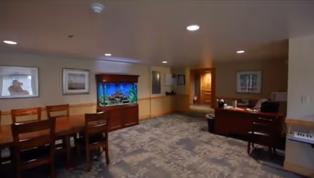 Interior view of a senior living community common area featuring a wooden dining table with chairs on the left, a large fish tank in the center against the wall, and a reception desk with office supplies on the right. The room has carpeted flooring with a floral pattern and framed pictures on the walls.