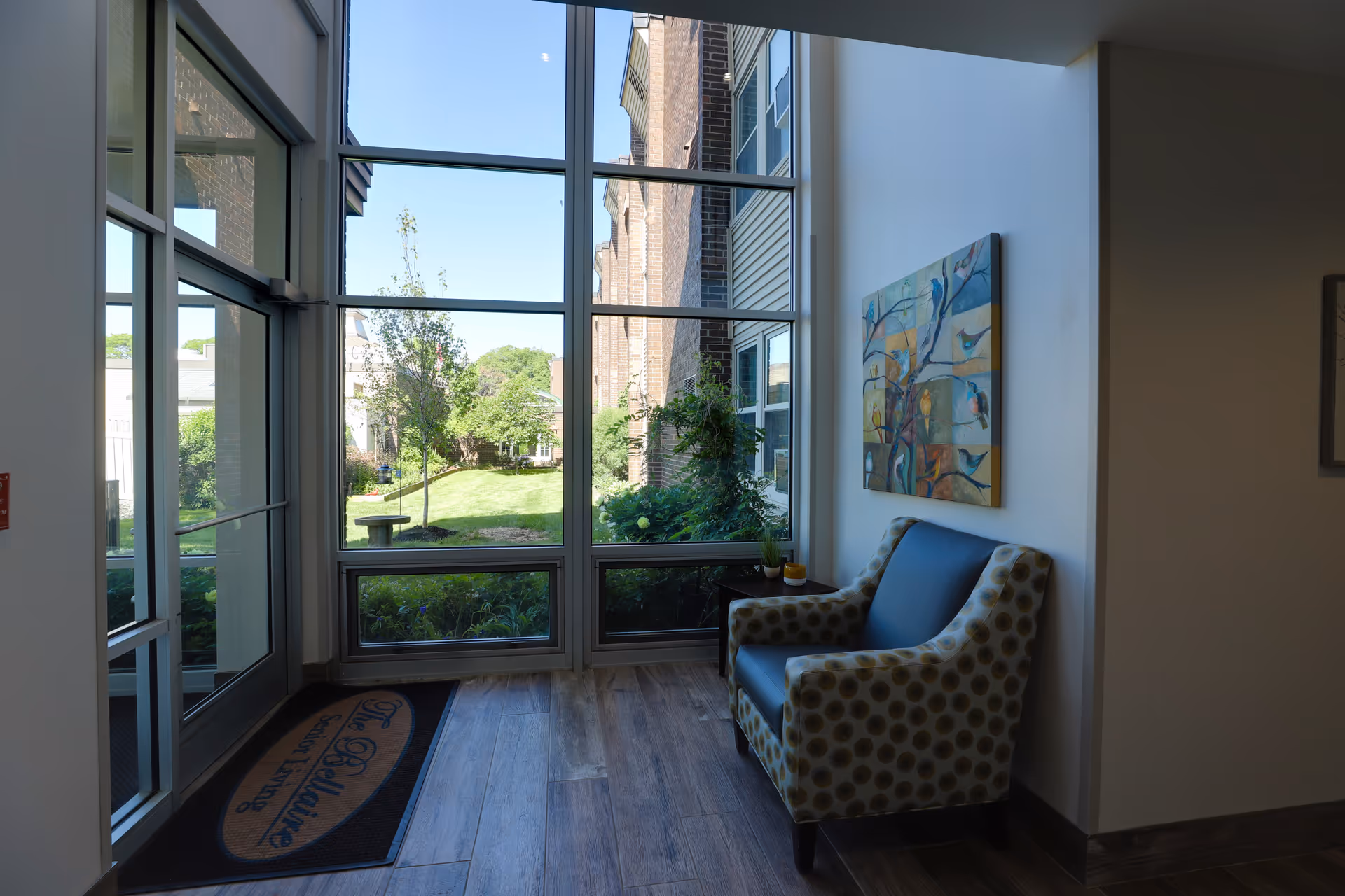 A bright seating area near a large window with a view of a green garden outside. The area features a patterned armchair with a blue cushion, a small side table with decorative items, and a colorful painting of birds on the wall. A door mat with the text 'The Bellaire Senior Living' is placed near the glass door.