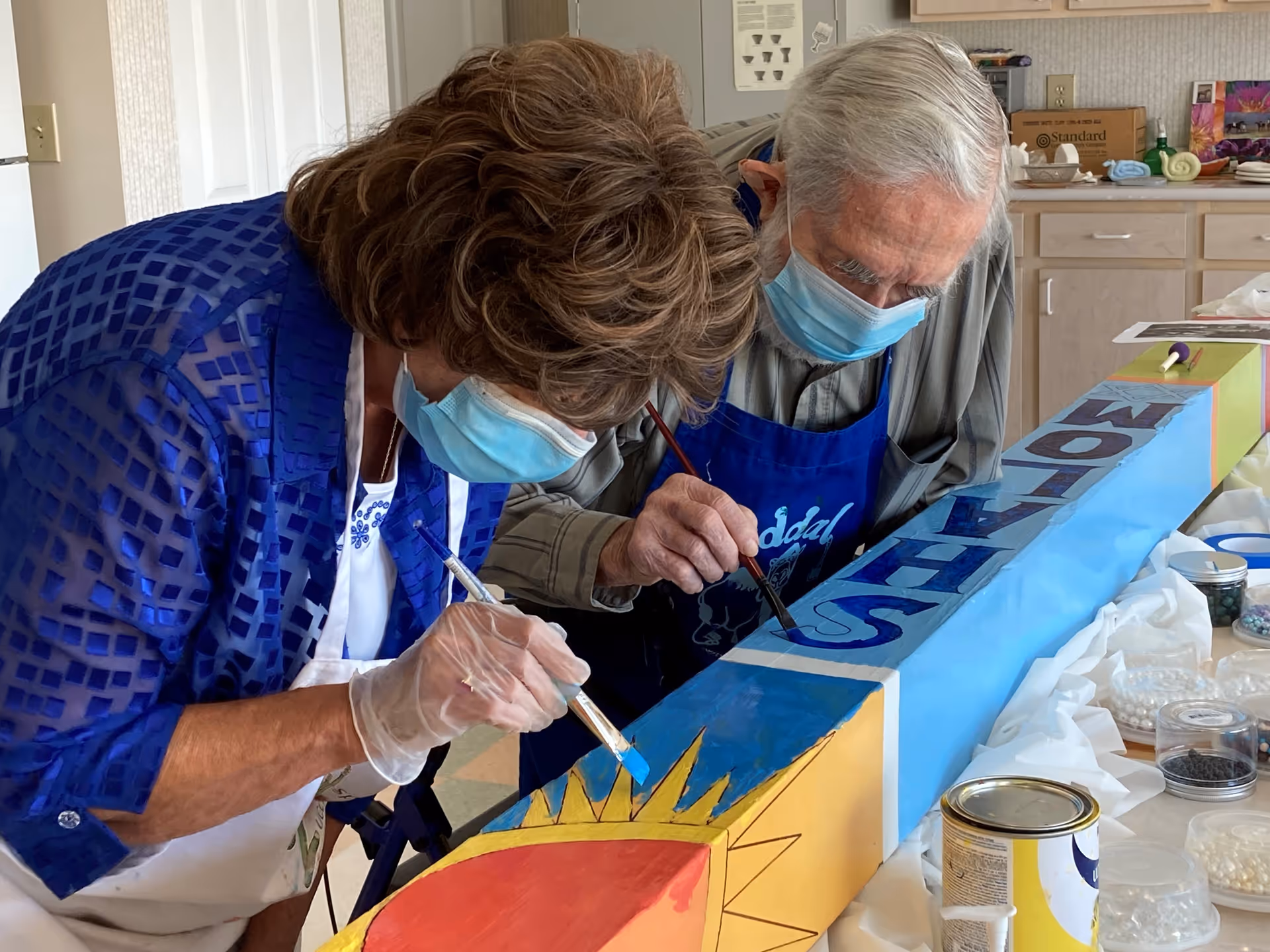 Two elderly individuals wearing face masks and aprons are painting a colorful wooden beam indoors. The beam features a sun design and the word 'MOTHS' in large letters. Various art supplies and containers are visible on the table around them.