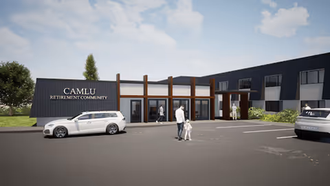 Exterior view of the CAMLU Retirement Community building with a parking lot in front. Several people are walking near the entrance, and cars are parked in the lot. The sky is partly cloudy.