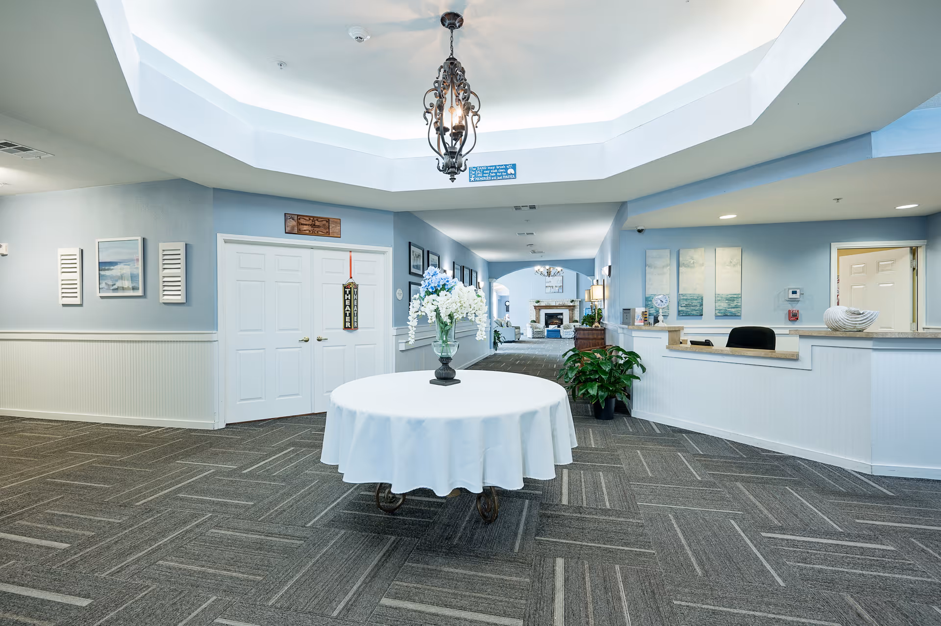 Interior view of a senior living facility lobby area with a round table covered with a white tablecloth and a vase of flowers in the center. The walls are painted light blue with white wainscoting, and there is a decorative chandelier hanging from the ceiling. A reception desk is visible on the right side, and a hallway leads to a room with a fireplace in the background. The space is well-lit and decorated with framed artwork and plants.
