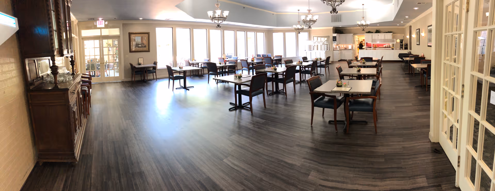 Spacious retirement community dining room with multiple tables and chairs, a buffet counter at the far end, chandeliers, and large windows.