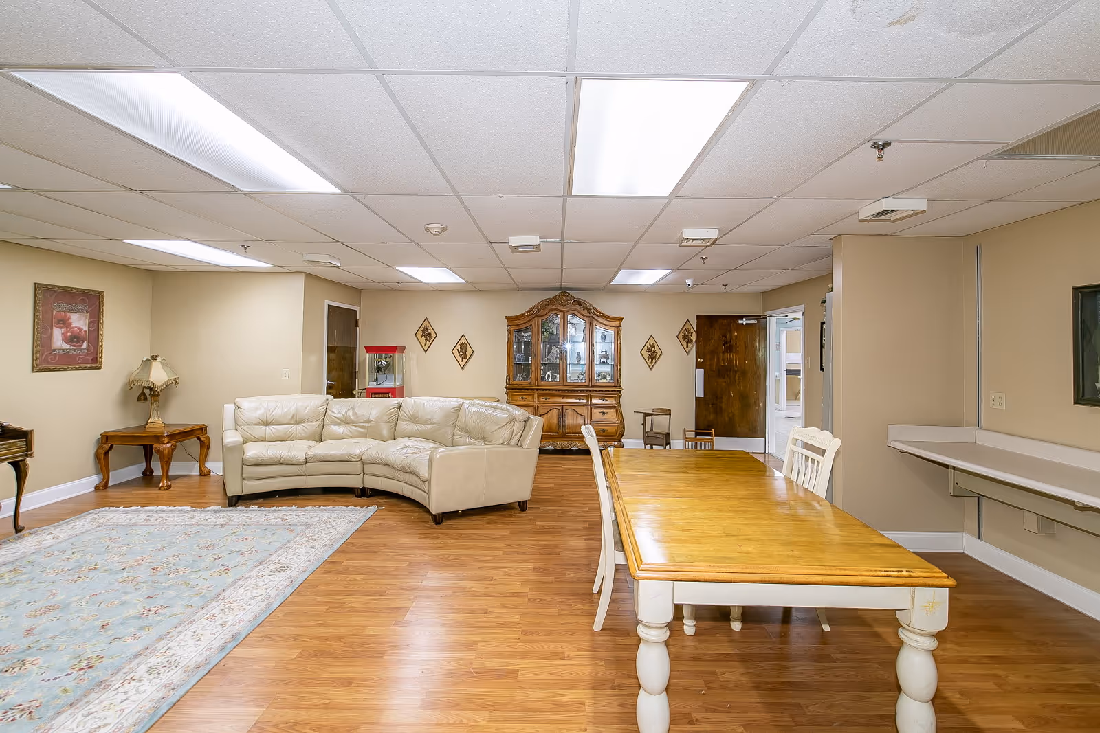 A spacious senior living common area with a beige sectional sofa, a wooden dining table with white chairs, a wooden cabinet with glass doors, a side table with a lamp, a framed picture on the wall, and a popcorn machine in the corner. The room has wood flooring, beige walls, and a drop ceiling with fluorescent lighting.