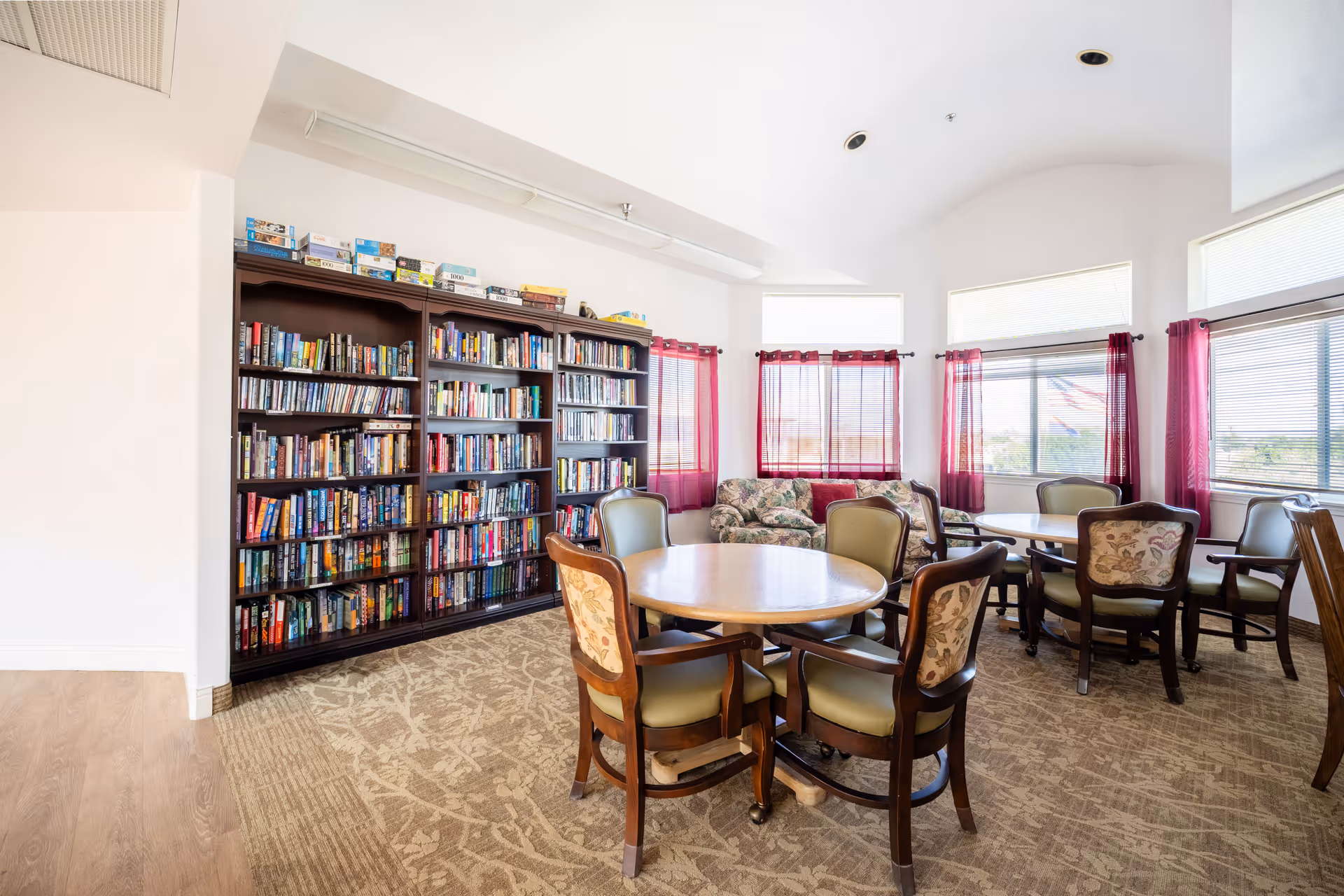 A bright and spacious room in Desert Rose Retirement Community featuring a large bookshelf filled with books and board games, round tables surrounded by cushioned chairs, and windows with red curtains letting in natural light. There is also a floral-patterned sofa near the windows.