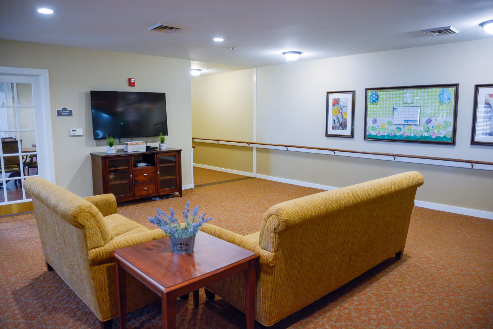 A cozy sitting area in a senior living facility with two mustard yellow sofas facing a wall-mounted flat-screen TV above a wooden cabinet. A small wooden table with a potted lavender plant is placed between the sofas. The walls are decorated with framed pictures and a bulletin board. The carpet is patterned in warm tones, and there is a hallway with handrails visible in the background.