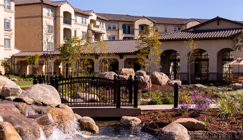 Courtyard featuring a small waterfall, pond, rock landscaping and a pedestrian bridge in front of a multi-story senior living building.