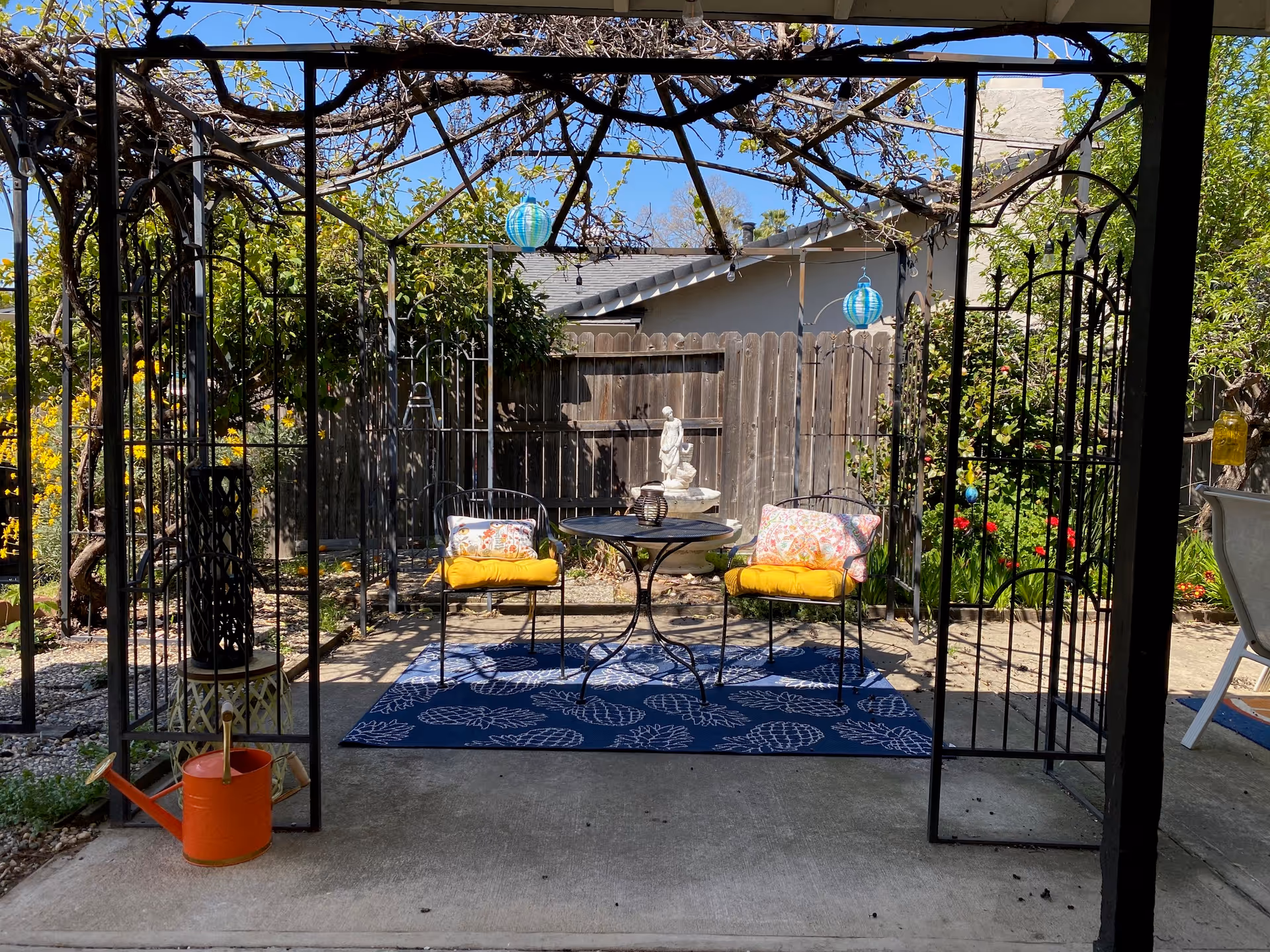 Outdoor patio area with a metal pergola covered in vines. Underneath, there is a small round black metal table with two matching chairs, each with yellow seat cushions and decorative pillows. A blue outdoor rug with white leaf patterns is placed under the table and chairs. In the background, there is a wooden fence, greenery, flowers, and a small statue on a pedestal. An orange watering can is on the ground to the left.