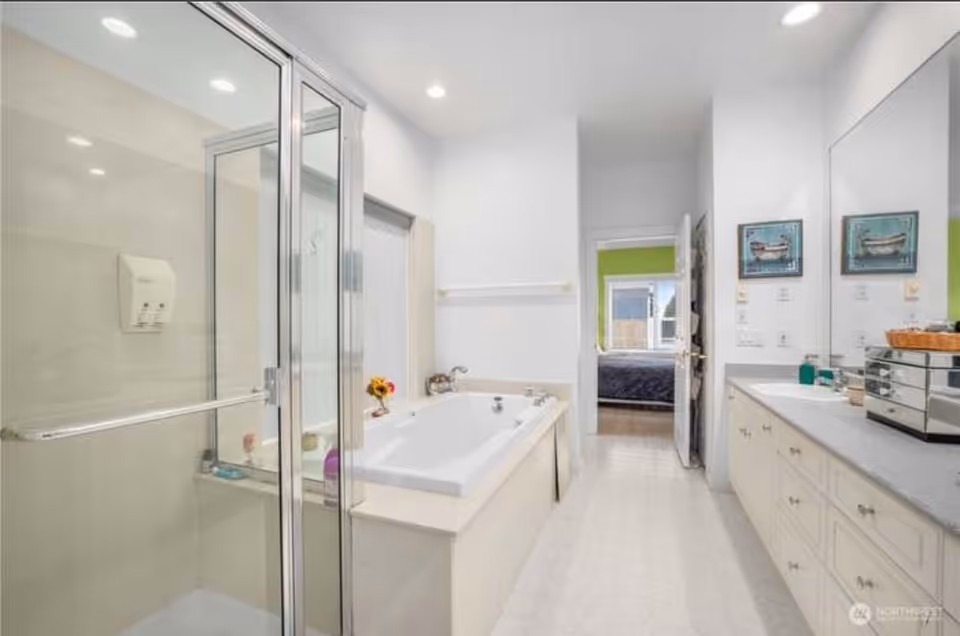 A bright, clean bathroom featuring a glass-enclosed shower on the left, a large bathtub in the center, and a long countertop with a sink and storage cabinets on the right. The bathroom has white walls and flooring, with two framed pictures above the counter and a doorway leading to a bedroom with a green accent wall.