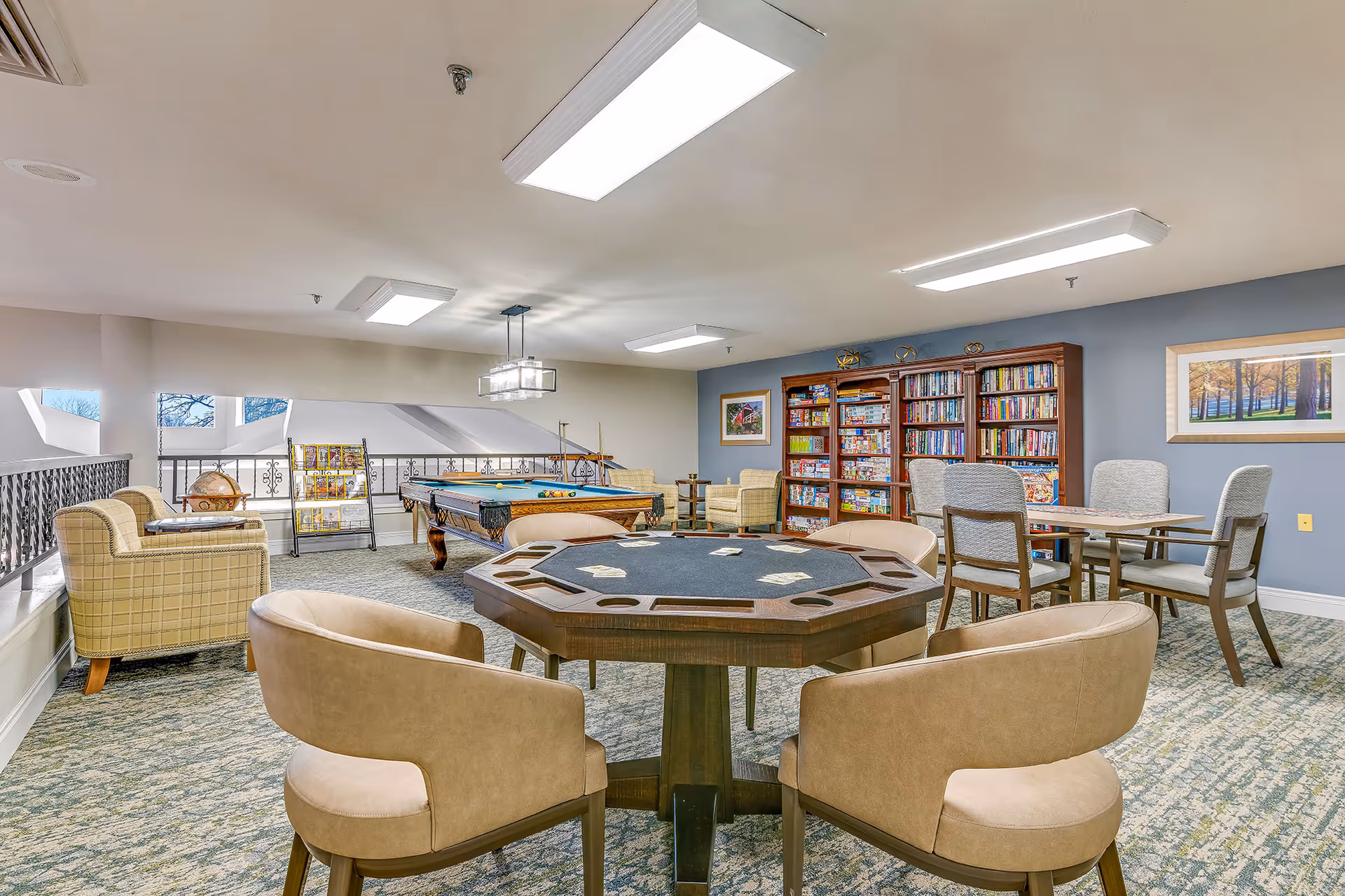 A senior living community game room featuring a card table with four beige chairs, a pool table, bookshelves filled with board games and books, additional seating with armchairs, and a table with four chairs. The room has carpeted floors, overhead fluorescent lighting, and framed artwork on the walls.