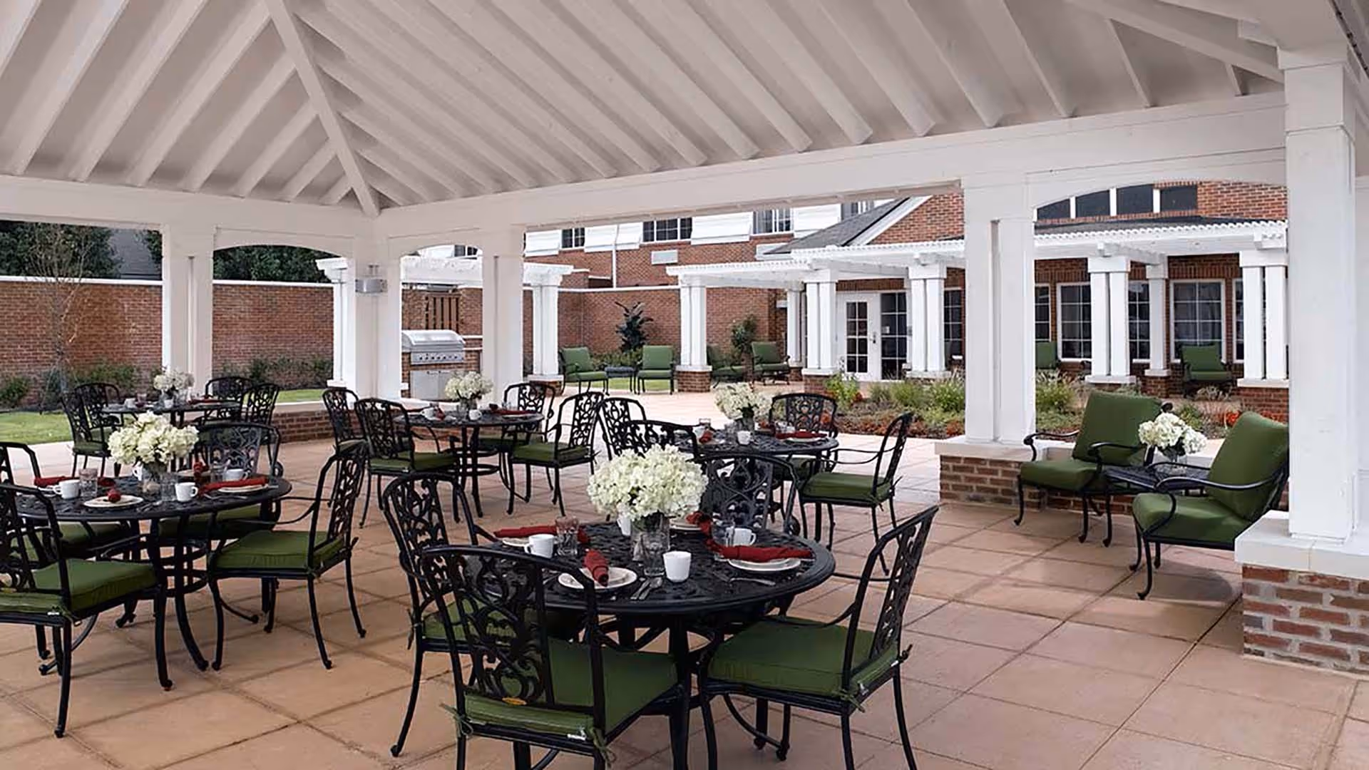 Covered outdoor patio area with black metal tables and chairs featuring green cushions. Each table is set with white flowers, plates, cups, and red napkins. The patio has a tiled floor and white wooden beams supporting the roof. In the background, there is a brick building with white columns and additional seating.