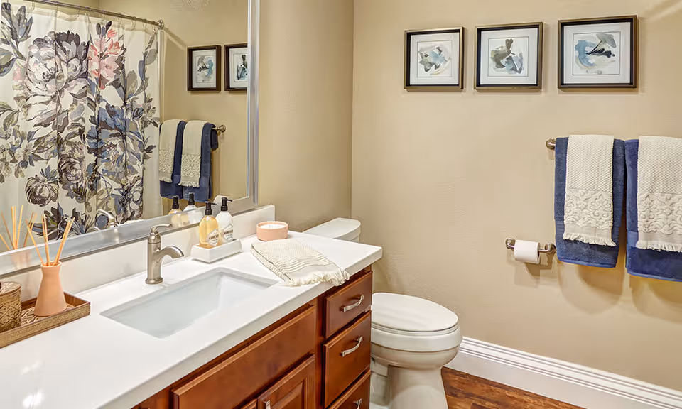 A clean and well-decorated bathroom featuring a white sink with a silver faucet set in a wooden vanity with drawers. On the countertop are soap dispensers, a small container, a folded towel, and a reed diffuser. A large mirror is mounted above the sink. The toilet is positioned next to the vanity, and above it are three framed abstract art pieces. Two blue and white towels hang on a towel rack beside the toilet, and a floral shower curtain is visible in the reflection of the mirror.