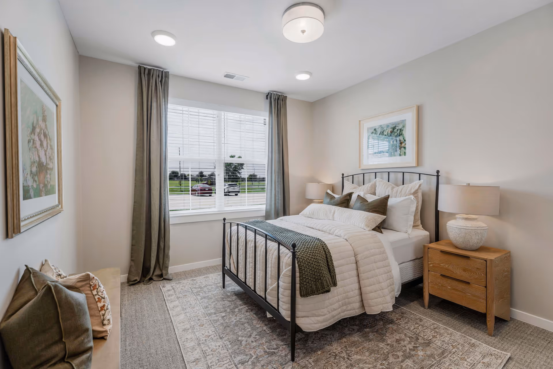 A cozy bedroom with a neatly made bed featuring white and green pillows and a green throw blanket. The room has a large window with gray curtains, a wooden nightstand with a lamp, a framed picture above the bed, and a bench with pillows along the left wall. The floor is carpeted with a patterned area rug.