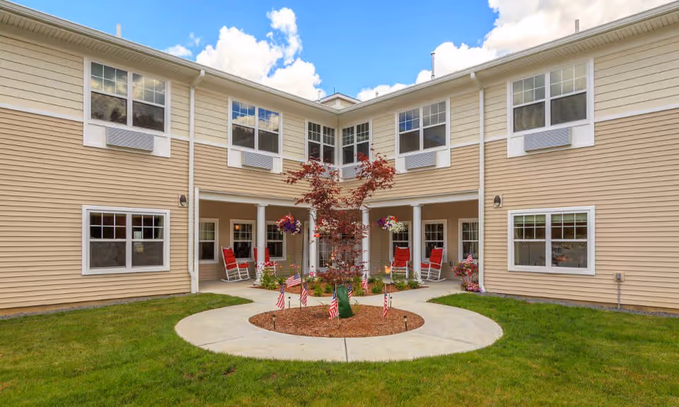 Outdoor courtyard area of a senior living facility with beige siding building surrounding a circular concrete walkway. In the center is a small tree with mulch and several small American flags. Red rocking chairs and hanging flower baskets are visible on the covered porch area. The sky is blue with some clouds.