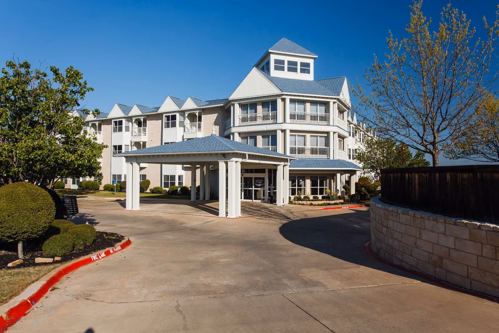 Front entrance and driveway of a multi-story white senior living building with a covered porte-cochere, landscaped bushes, and a clear blue sky.