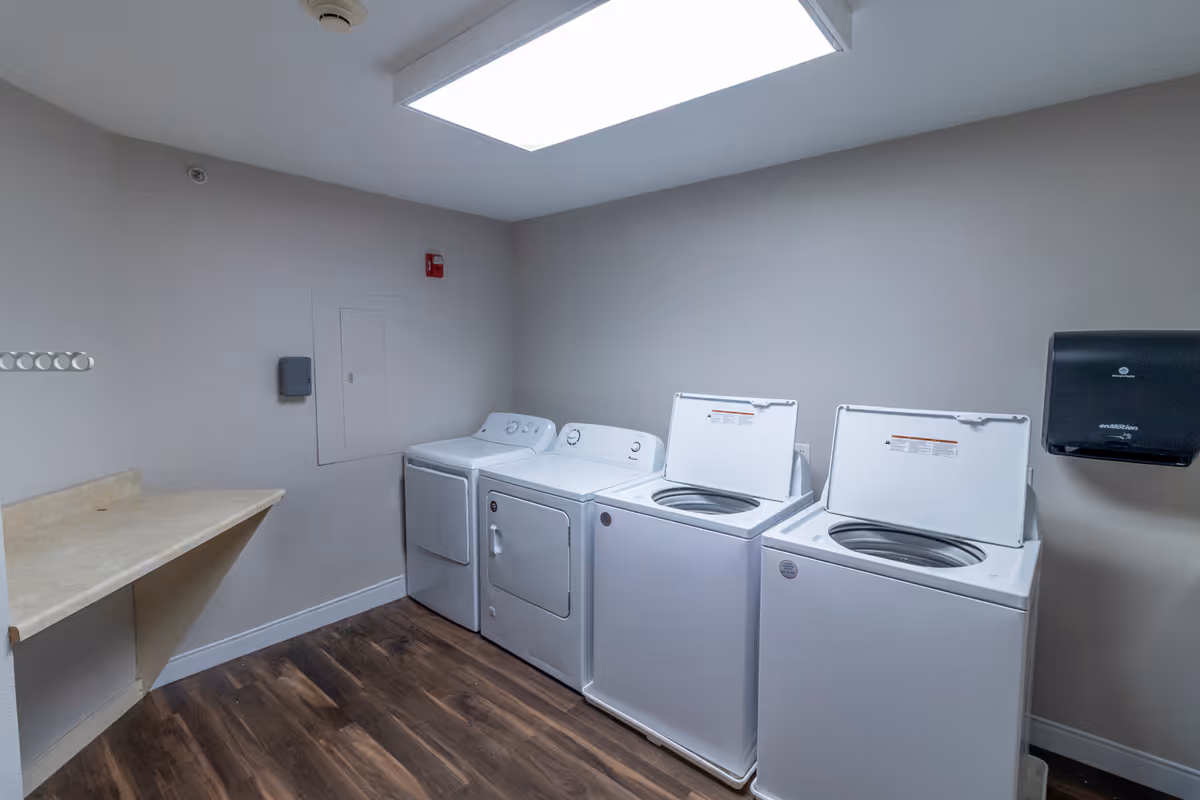 Laundry room with two top-loading washing machines and two front-loading dryers lined up against a beige wall. There is a countertop on the left side and a paper towel dispenser mounted on the right wall. The room has wood-look flooring and a large rectangular ceiling light.