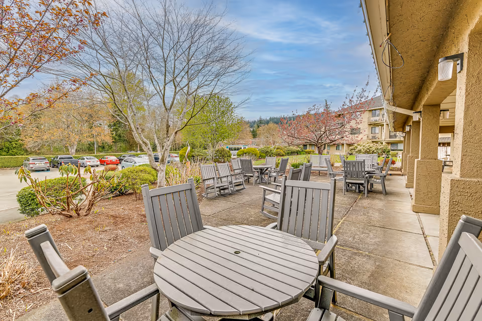 Outdoor patio at a senior living facility with round wooden tables and chairs on a concrete terrace, landscaped beds and trees, and parked cars in the background.