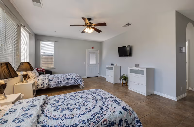 Spacious bedroom with two twin beds covered in blue patterned quilts, bedside lamps, a wall-mounted TV, ceiling fan, large windows, and tile flooring.