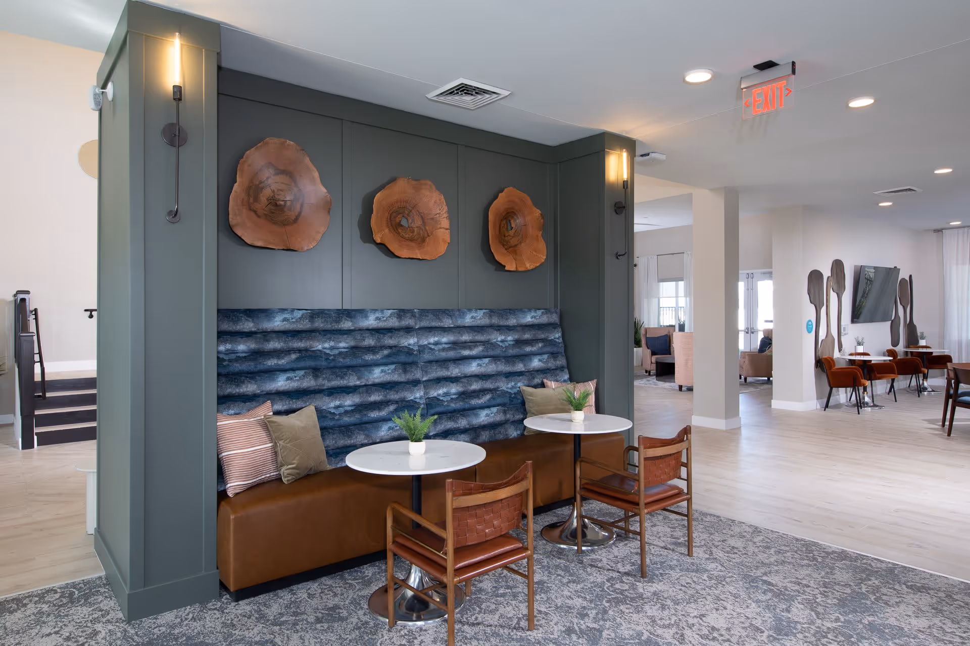 A modern seating area in a senior living facility with a blue cushioned bench against a green wall adorned with three wooden art pieces. Two small round tables with potted plants and two brown leather chairs are positioned in front of the bench. The space has light wood flooring, a patterned rug, and is well-lit with ceiling lights and wall sconces. In the background, there are additional seating areas with chairs and a wall-mounted TV.