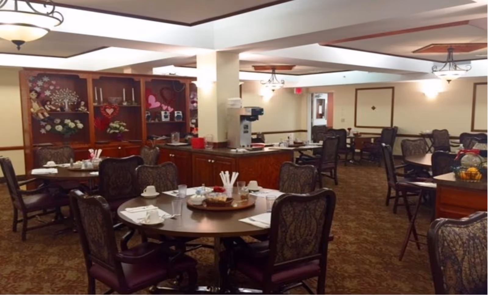 A dining room in a senior living facility with round tables set with cups, glasses, napkins, and utensils. The room features upholstered chairs, a carpeted floor, and wooden cabinetry with decorative items including flowers and heart-shaped decorations. Ceiling lights provide warm lighting.
