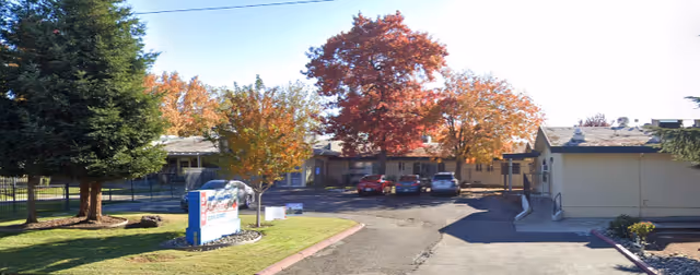 Exterior view of North Pointe Care Center showing a driveway leading to the building entrance with several parked cars. Trees with autumn-colored leaves surround the area, and a sign is visible near the driveway entrance.
