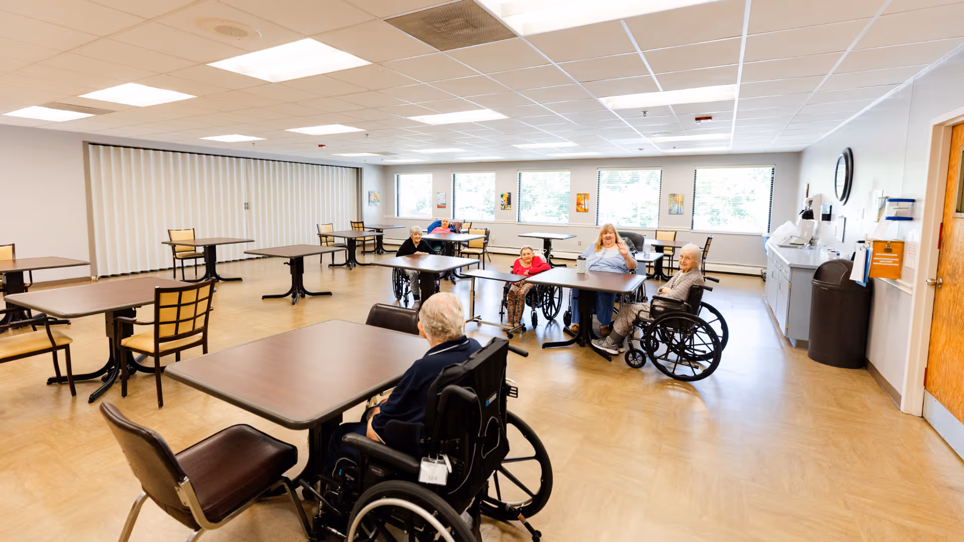 A spacious, well-lit common room in a healthcare facility with several elderly individuals sitting around tables, some in wheelchairs. The room has large windows letting in natural light, multiple tables and chairs, and a clean, organized appearance.