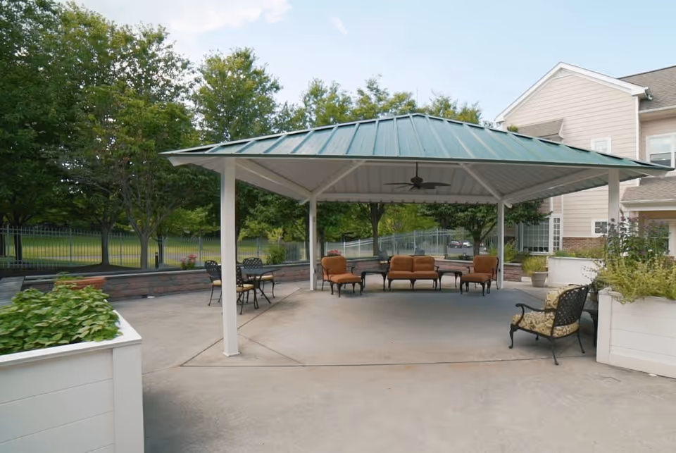 Covered outdoor pavilion with seating and tables on a concrete patio next to a residential building and landscaped grounds.