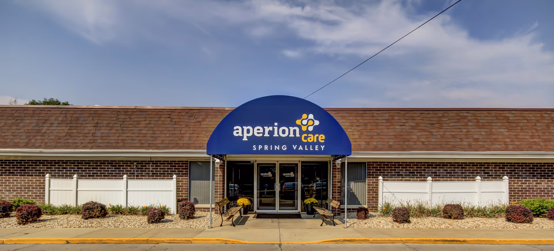 Front entrance of Aperion Care Spring Valley in a single-story brick building with a blue arched awning and glass double doors.
