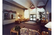 A spacious living room area with high vaulted ceilings and large windows covered with sheer curtains. The room is furnished with a floral-patterned sofa, wooden tables, lamps, a potted plant, and a dining table with chairs near the back. The carpet is dark, and the walls are light-colored, creating a warm and inviting atmosphere.