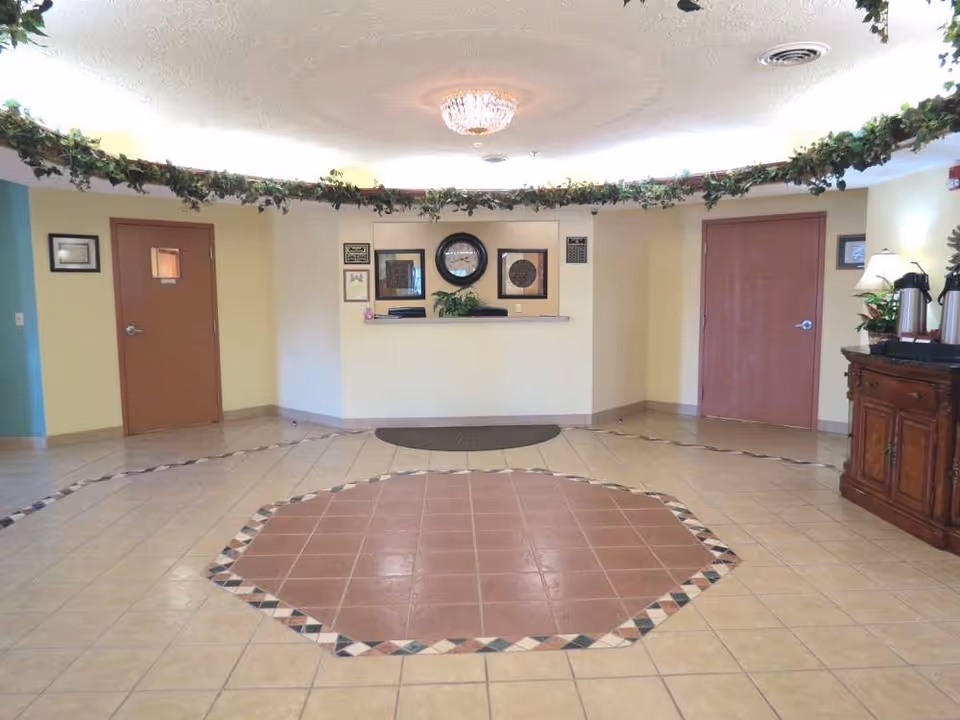 Reception area with a tiled floor featuring a central octagonal pattern, a white reception desk with framed pictures and a clock on the wall behind it, two closed wooden doors on either side, a wooden cabinet with coffee dispensers and a lamp on the right, and greenery decoration along the ceiling edge.