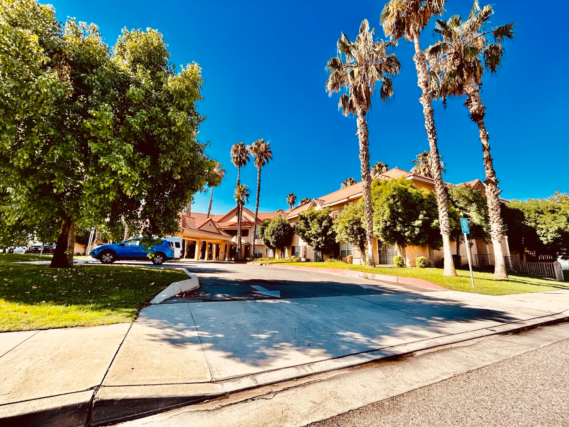 Exterior view of a senior living facility with a driveway leading to the entrance. The building has a red-tiled roof and is surrounded by tall palm trees and other green trees under a clear blue sky. A blue car is parked near the entrance.