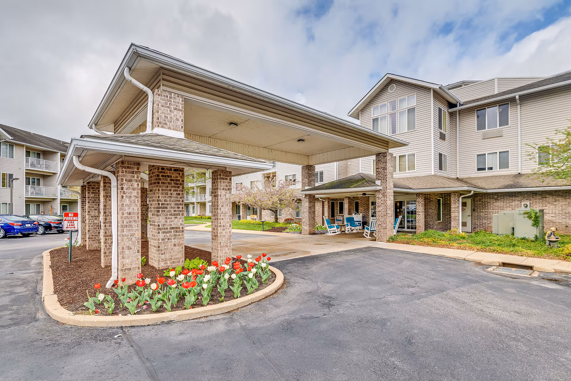 Entrance and porte-cochere of a multi-story senior living building with brick columns, planted tulips, and a driveway.