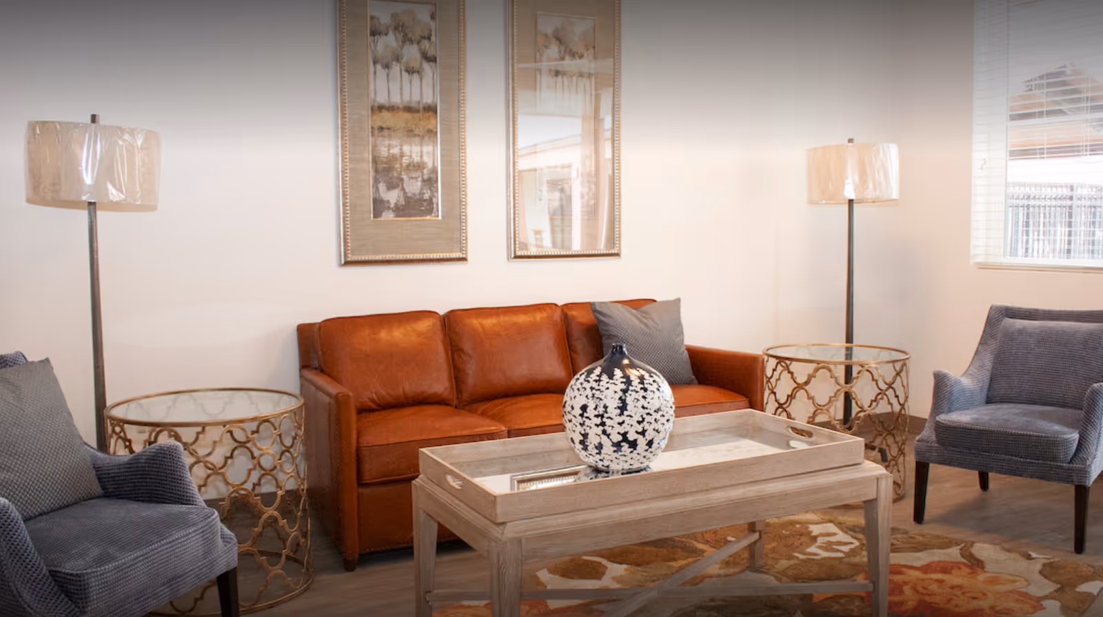 A cozy living room with a brown leather sofa, two gray upholstered armchairs, two round gold side tables with glass tops, two floor lamps with beige shades, a wooden coffee table with a decorative black and white vase, and two framed artworks on the wall.