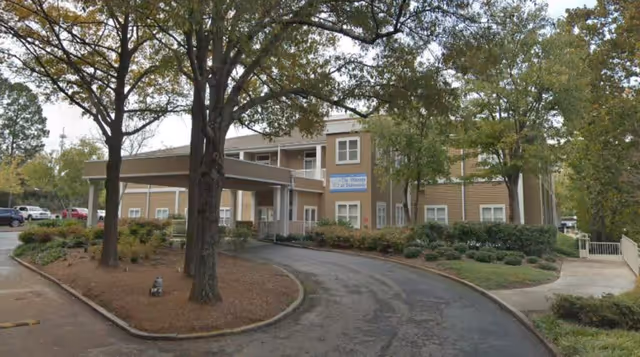 Exterior view of The Phoenix at Dunwoody senior living facility showing a two-story beige building with white trim, a covered entrance driveway, trees, and landscaped bushes.