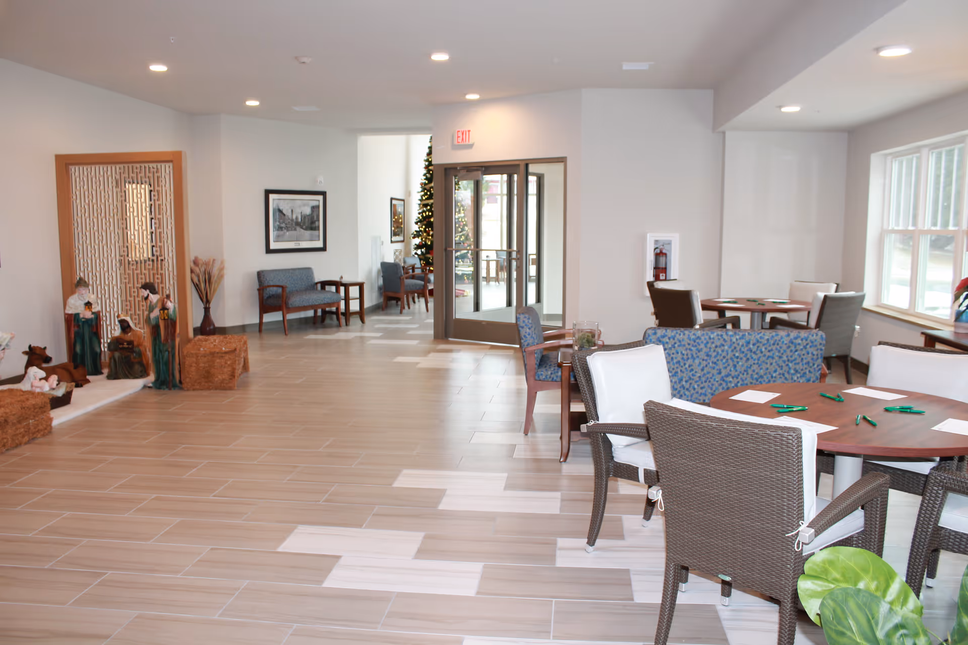 A bright and spacious common area in a senior living facility with several round tables and chairs arranged for seating. There are large windows on the right side letting in natural light. In the background, there is a glass door entrance, a Christmas tree, and some seating benches along the wall. On the left side, there is a nativity scene display with figurines and decorative elements.