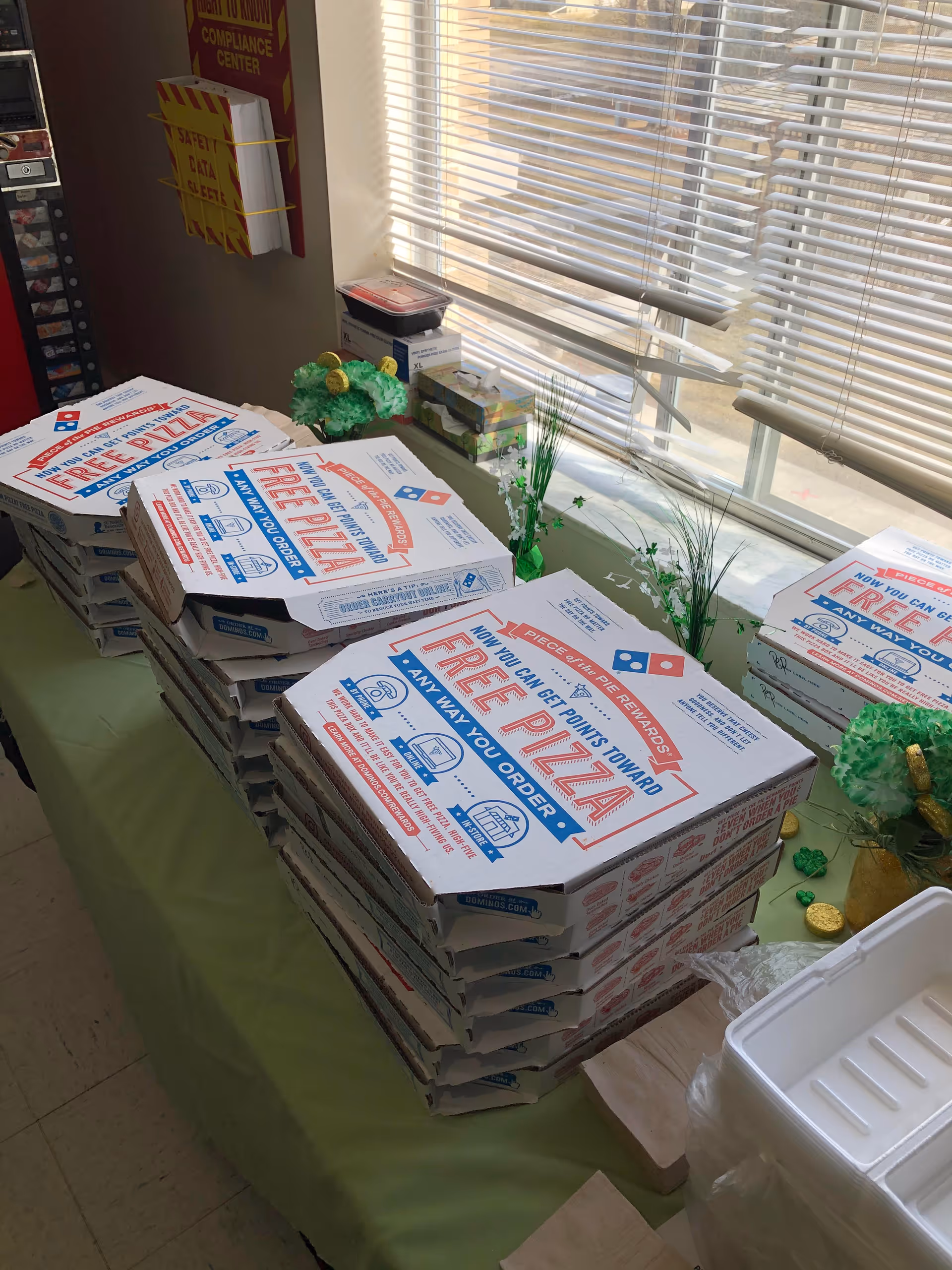 A table covered with a green tablecloth holding multiple stacked Domino's pizza boxes near a window with blinds. There are some small decorative plants and a few other items on the table and windowsill.