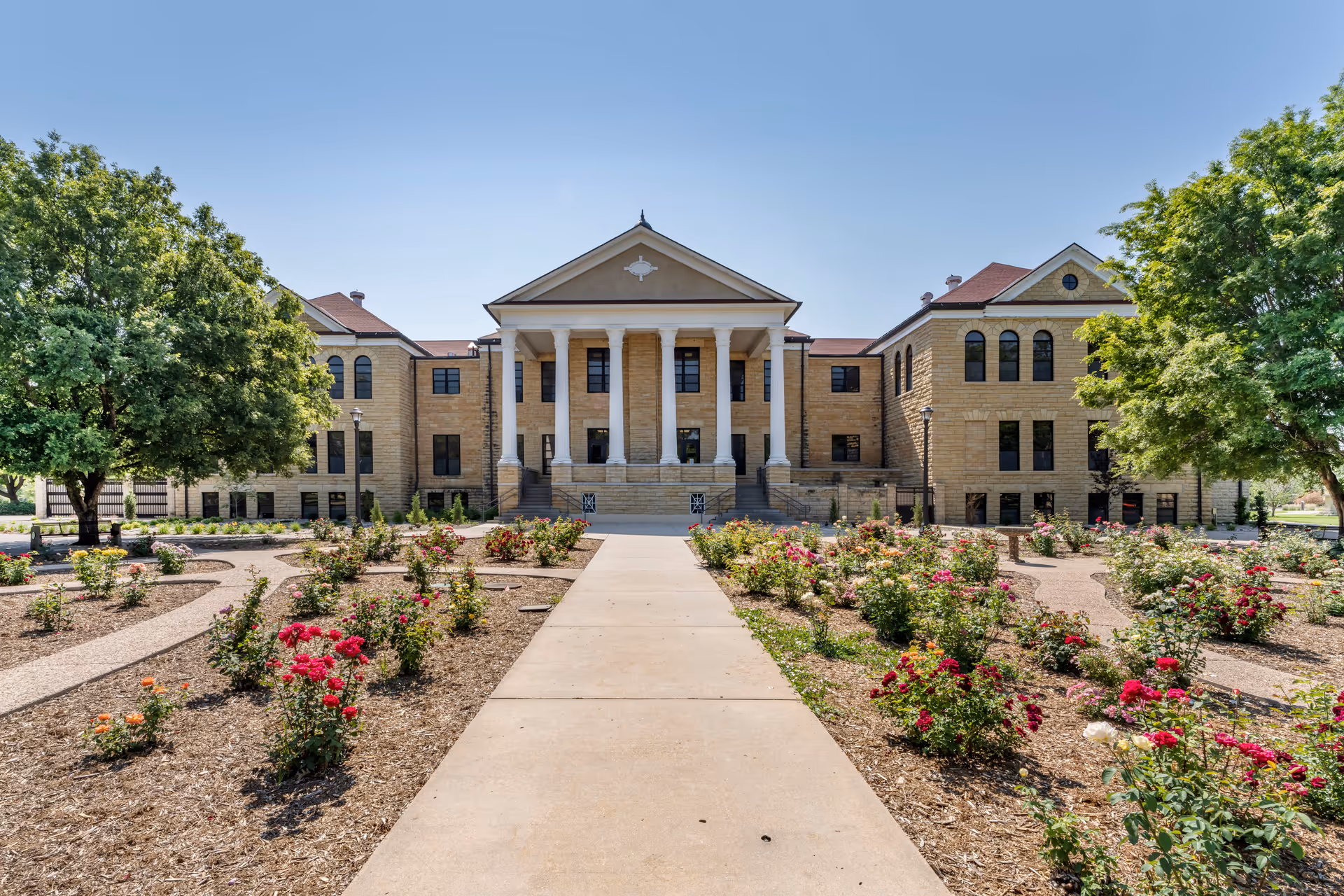 Front exterior view of a large, historic-style building with beige stone walls and a red roof. The building features a prominent entrance with tall white columns and steps leading up to it. In the foreground, there is a garden with neatly arranged flower beds and a concrete pathway leading to the entrance. Two large green trees flank the garden on either side under a clear blue sky.