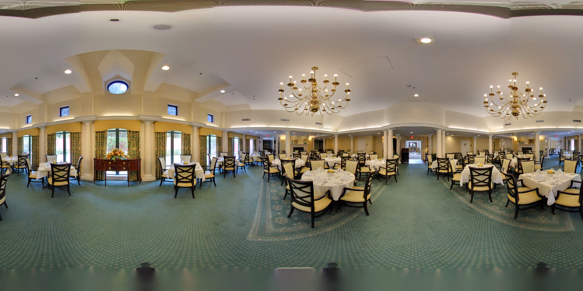 A spacious, elegant dining room with multiple round tables covered with white tablecloths and set with plates, glasses, and napkins. The room features green carpet with a patterned border, large windows with curtains, cream-colored walls with columns, and two ornate chandeliers hanging from the ceiling. A wooden sideboard with a floral arrangement is positioned against one wall.