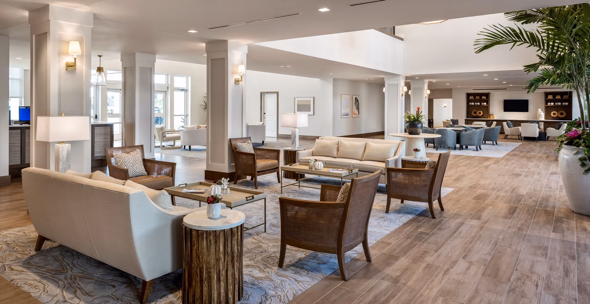 Spacious and well-lit senior living facility common area with multiple seating arrangements including sofas, armchairs, coffee tables, and side tables. The room features wooden flooring, large potted plants, wall sconces, and framed artwork on the walls. There is a reception desk visible in the background and a television mounted on the far wall.